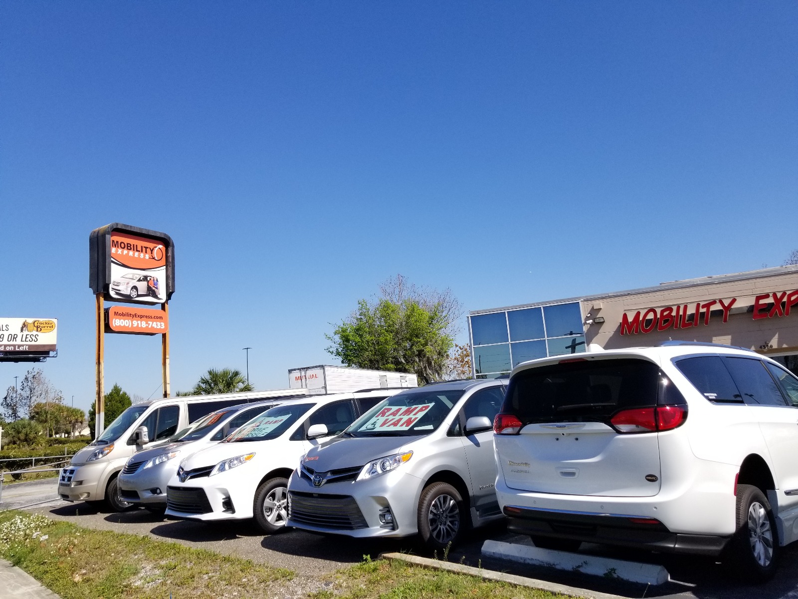 A line of handicap accessible vans parked with most facing forward, in front of a building for the wheelchair van dealer Mobility Express. There's a tall orange colored sign in the background to the left that also has the words "Mobility Express" on it.