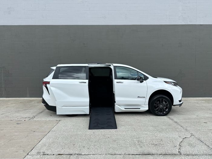 Facing the passenger side of a white Toyota Sienna minivan. The passenger side sliding door is open, and a wheelchair ramp is deployed down to the ground. The background is a white and gray block wall.