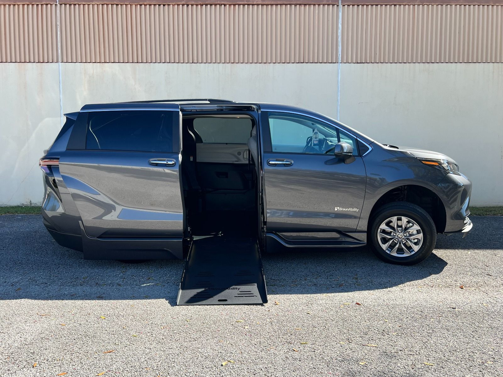 Facing the passenger side of a gray Toyota Sienna minivan. The passenger side sliding door is open, and a wheelchair ramp is deployed down to the ground. The background is a nondescript white wall with a rust colored corrugated metal roof.
