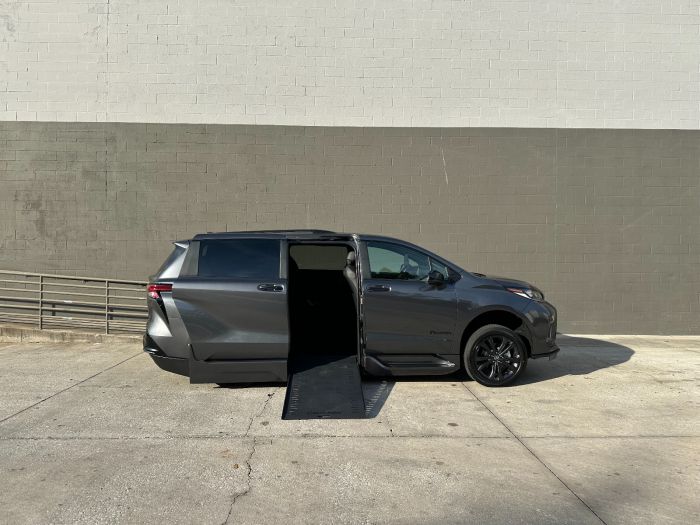 Facing the passenger side of a gray Toyota Sienna minivan. The passenger side sliding door is open, and a wheelchair ramp is deployed down to the ground. The background is a white and gray block wall.
