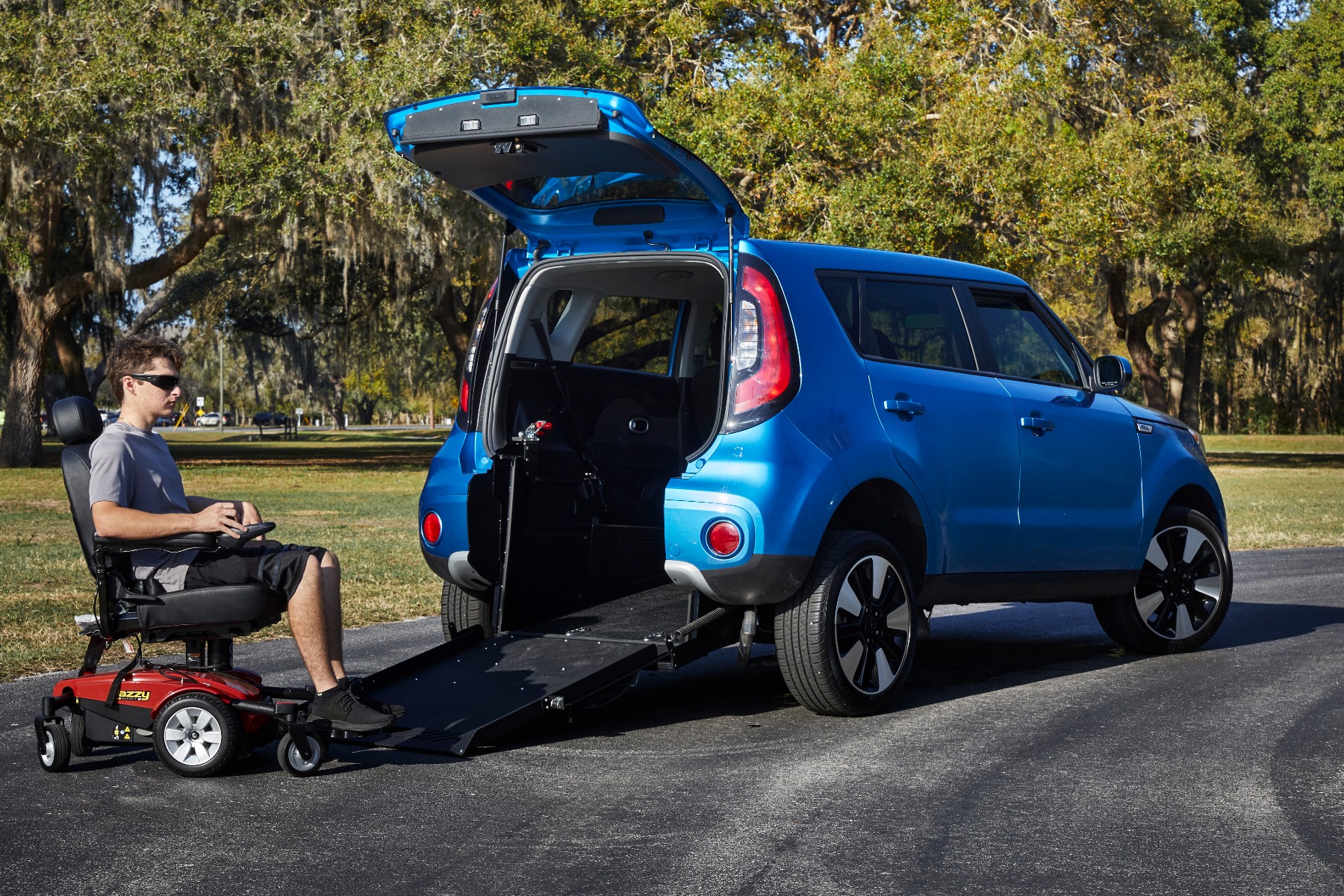 Preparing for a wheelchair van demonstration. A man is sitting on a red power wheelchair. The power wheelchair is at the bottom edge of a ramp deployed from the rear hatch of a wheelchair accessible Kia Soul small SUV. The Kia Soul is blue. The setting seems to be a park with trees and a road in the background.