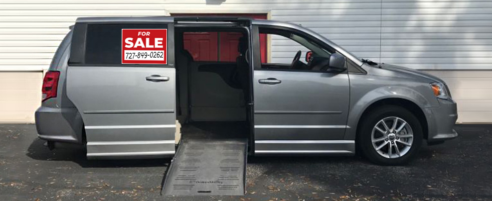 A silver handicap accessible minivan is parked on front of a white building. The passenger sliding door is open and there is a ramp deployed for a wheelchair to use.